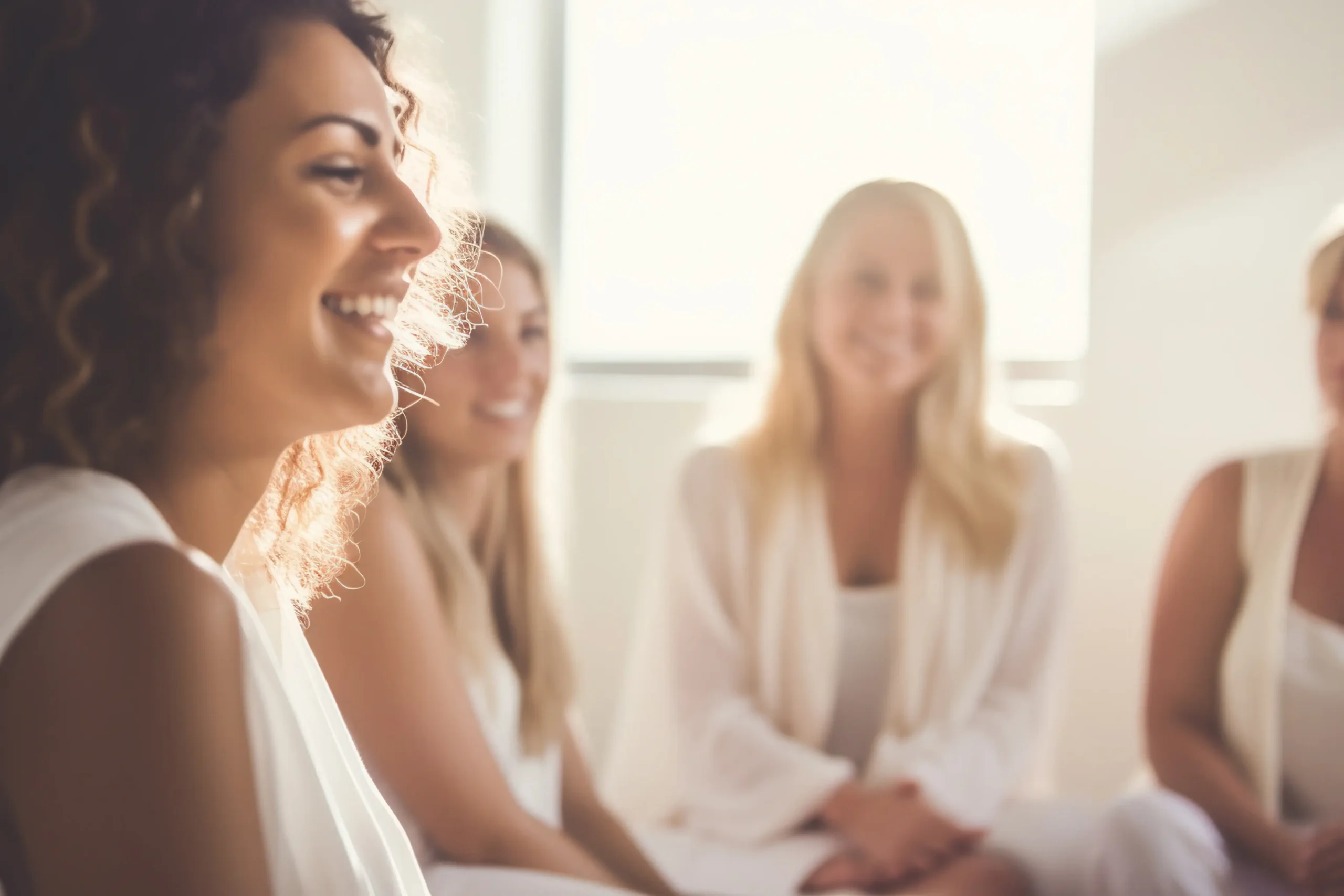 A group of women, dressed in white engaged in a thoughtful discussion in a bright softly lit room